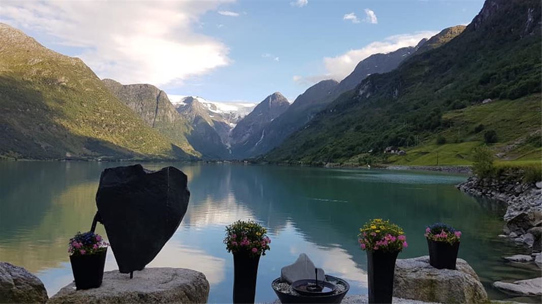 Calm Oldenvatnet lake reflects a valley and snowy peaks, with flower pots and a dark heart-shaped sculpture in front at Yrineset in Olden..
