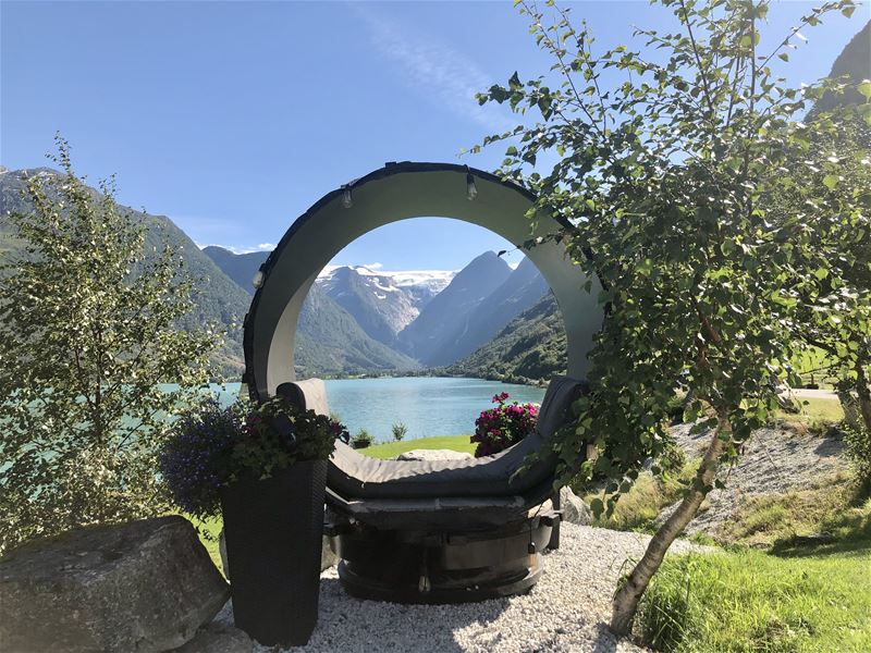 Circular outdoor lounge pod overlooks a turquoise lake and steep mountains, framed by trees under a clear blue sky..