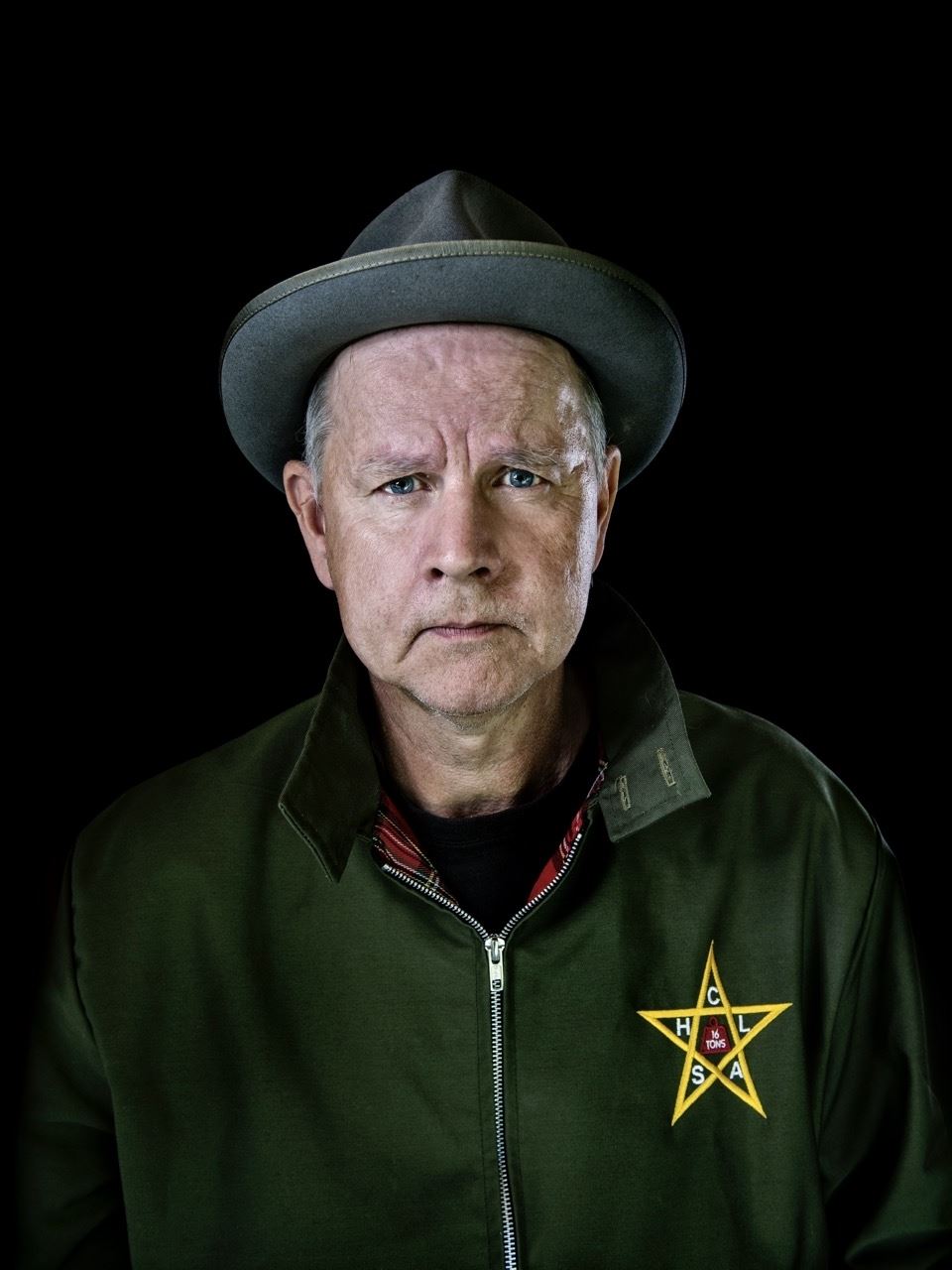 En man med en grå hatt på huvudetiklädd en grön jacka med en gul stjärna på bröstet, svart bakgrund.