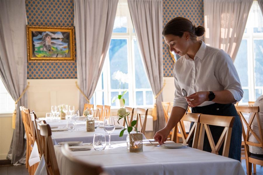 Ein servitør i kvit skjorte dekkjer eit bord med kvit duk, glas og blome, i ein lys restaurant med vindauge..
