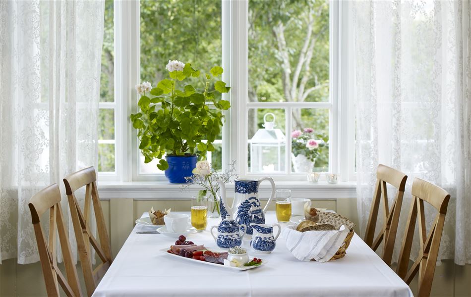 Dekka frukostbord med kvit duk, blå servise og brød, framfor stort vindauge med blondegardiner og grøne plantar..