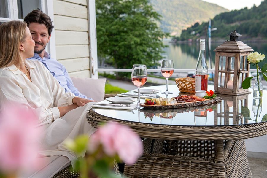 Eit par sit på ei terrasse med dekka bord, rosévin og snackfat, med fjord og skogkledde fjell i bakgrunnen..