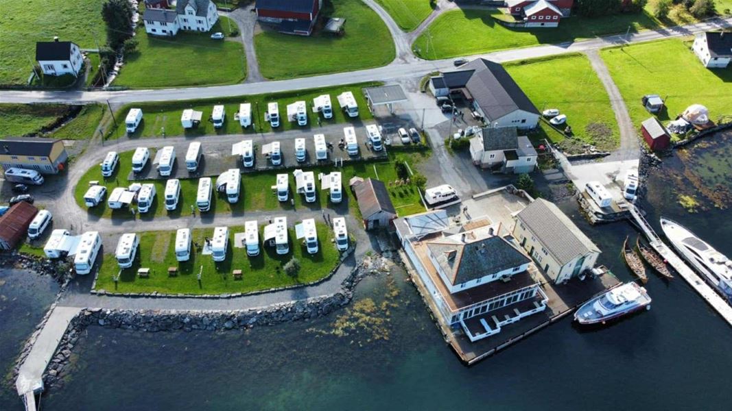 Aerial view over Davik Fjordparadis in Bremanger of a seaside RV campground with rows of white campers, nearby buildings, roads, and docked boats..