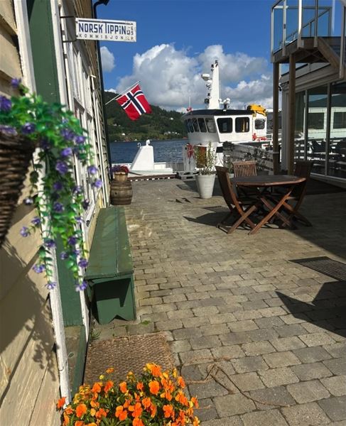 Sunny waterfront patio with wooden table and chairs, potted flowers, a docked boat, and a Norwegian flag..