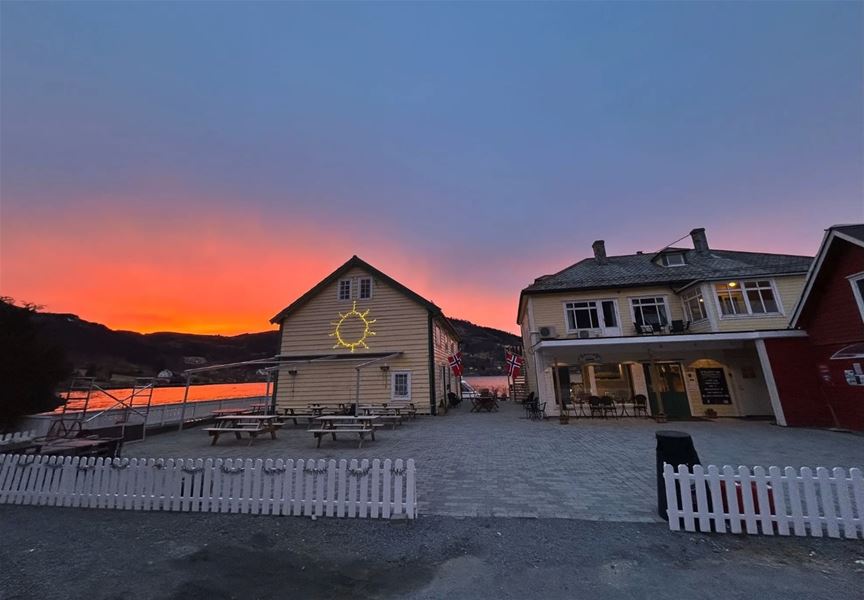 Two wooden buildings by a waterfront with picnic tables and a white fence under a vivid orange sunset sky at Davik Fjordparadis in Bremanger..