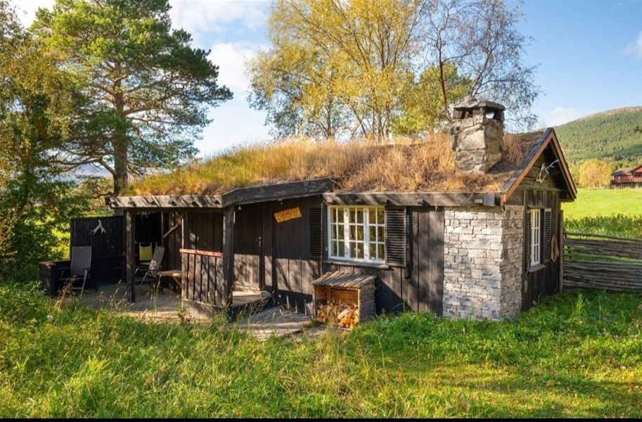 Utebilde av en tømmerbygning med stråtak omgitt av gress og trær, med fjellandskap i bakgrunnen.
