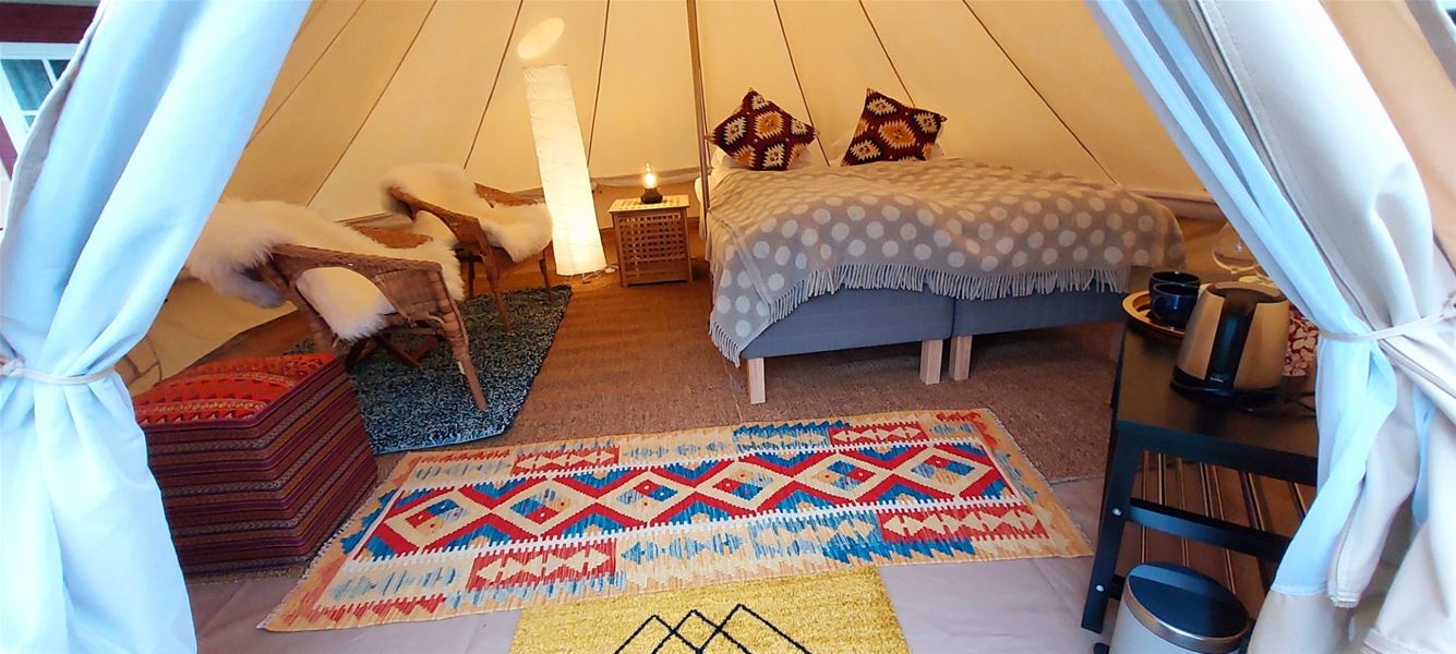 Innside av et glampingtelt med seng, tepper og en liten sitteplass, dekorert med varme farger og tekstiler.