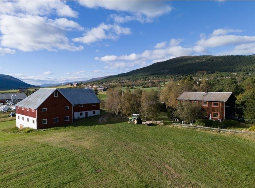 Luftfoto av tradisjonelle gårdsbygninger plassert i et åpent dalføre med jorder, trær og fjell i bakgrunnen under klar himmel.