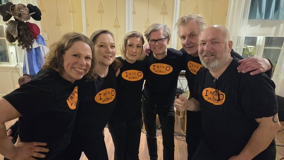 Fyra kvinnor och två män iklädda svarta t-shirts med orange text, de står i en halvcirkel och håller armarna på varandras axlar.