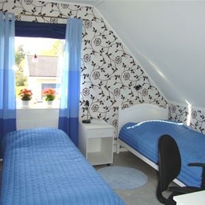 Två enkelsängar i ett rum med blå gardiner och svart- och vitblommig tapet.