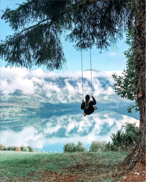 A person on a rope swing hangs from a tree above a grassy slope, overlooking a cloudy mountain lake at Skarsteinssætra i Olden.