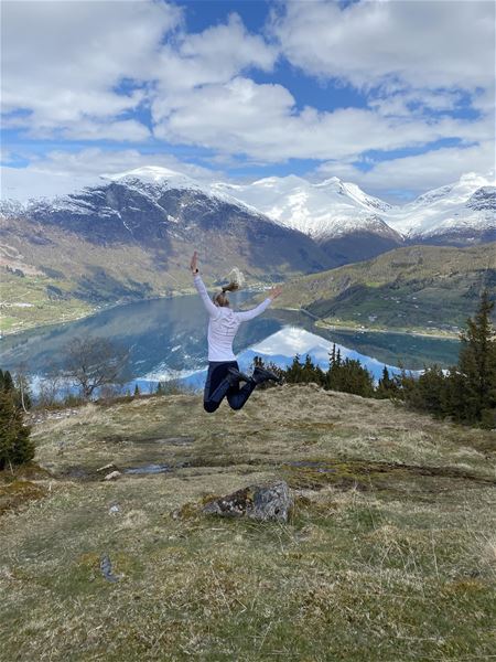 Person mid-jump on a grassy hill overlooking a blue fjord, forested slopes, and snow-capped mountains under clouds at Skarsteinssætra in Olden.
