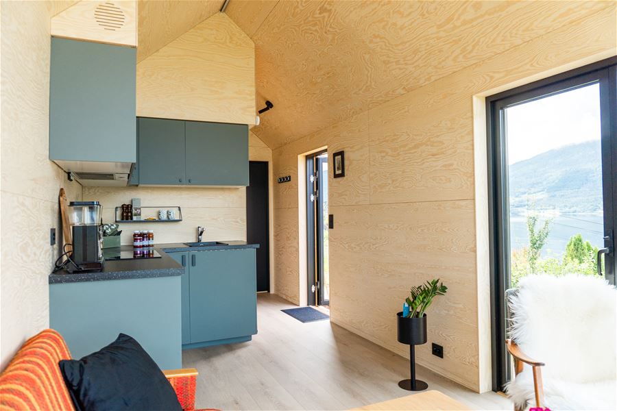 Interiør med lyse treveggar, blå kjøkeninnreiing, liten benk og vindauge med utsikt mot fjord og fjell..