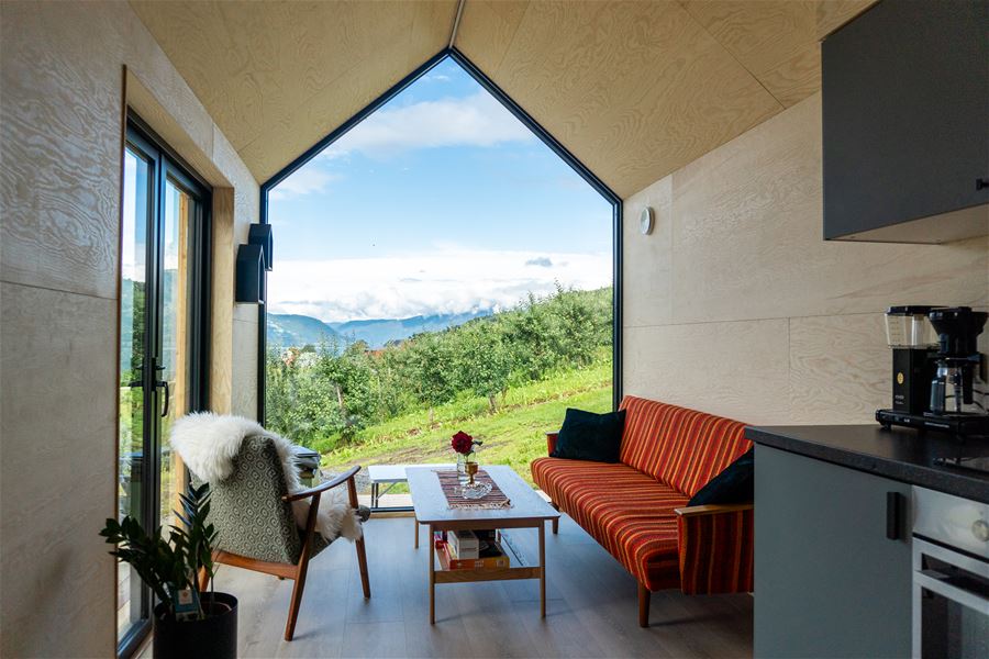 Moderne stoverom med sofa, lenestol og bord, og stor vindaugsopning mot grøne bakkar og fjell under blå himmel..