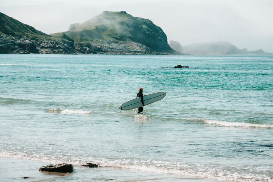 Ein surfear i Ervik i våtdrakt vassar i grunt hav med surfebrett, med klipper og tåkelagde fjell i bakgrunnen..