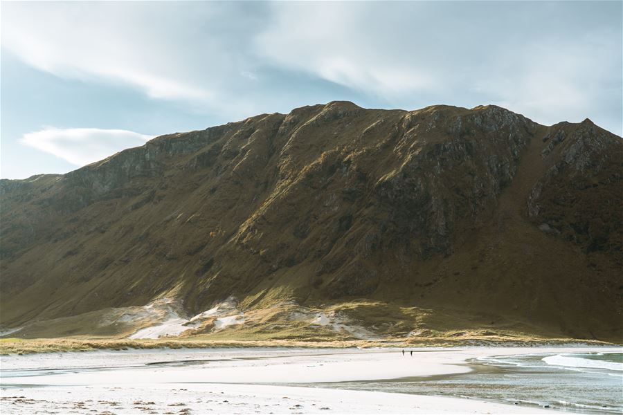 Stor fjellrygg reiser seg over ei lys sandstrand i Ervik med grunt vatn, under lettskyet himmel i mjukt lys..