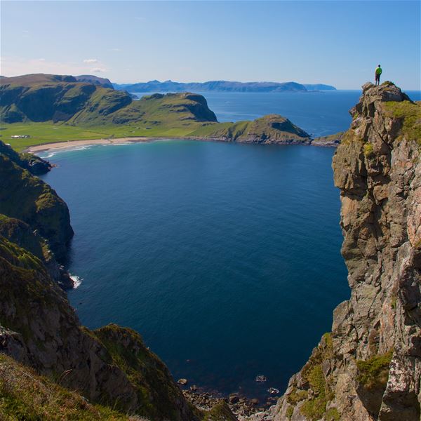 Ein person i grøn jakke står på ei høg klippe i Ervik og ser utover ei blå havbukt med fjell og strand..