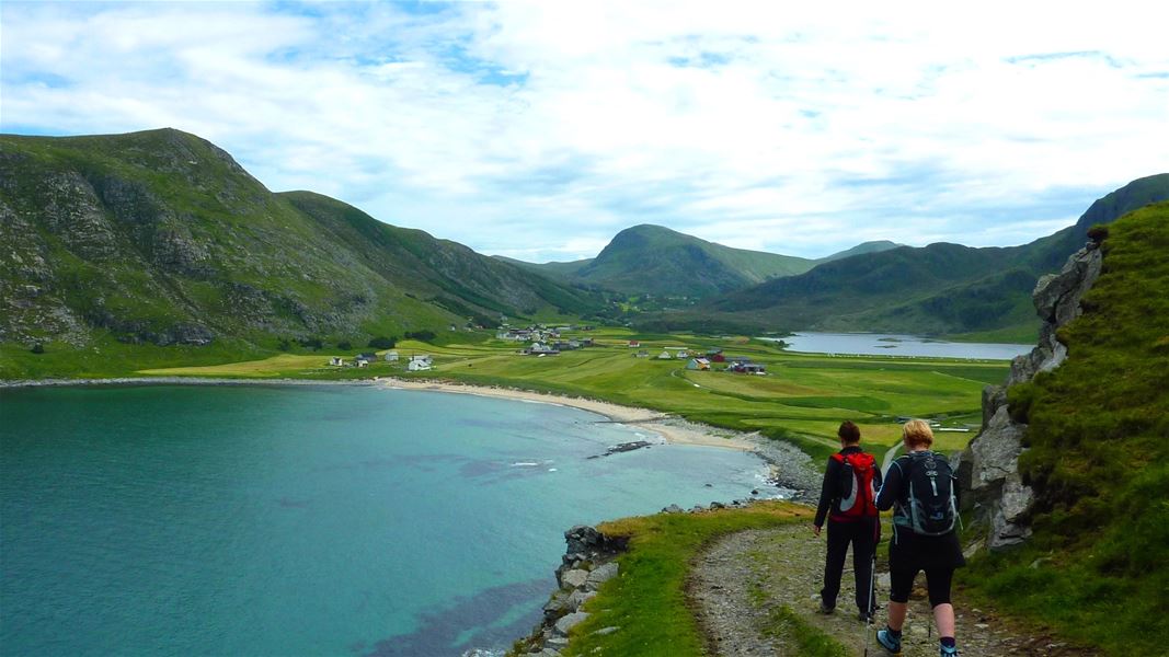 To turgåarar med ryggsekk går på ein sti over ein fjord, med grøne fjell, strand og bygder i bakgrunnen..