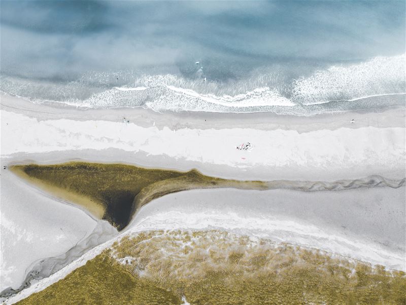 Luftfoto av bølgjer som slår mot ein lys sandstrand, med eit mørkare elvelaup og grasområde i framkant..