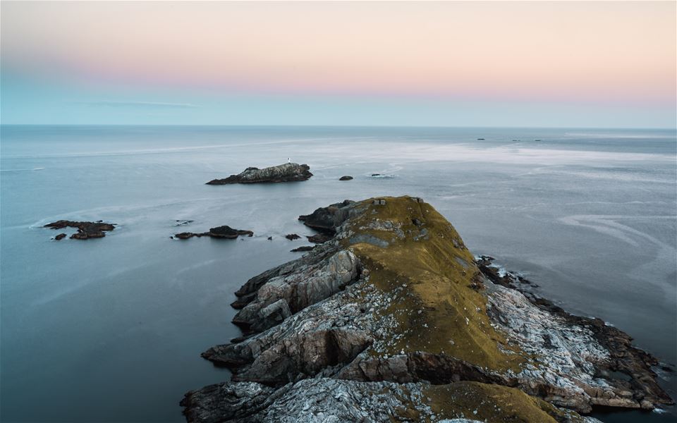 Luftfoto av ei smal, steinete holme med grasrygg i roleg hav under pastellfarga himmel ved skumring..
