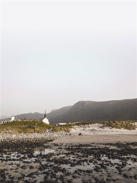 Kvit kyrkje og gravplass på ei grøn høgd ved ein steinete fjøre, med fjell i bakgrunnen og lys himmel..