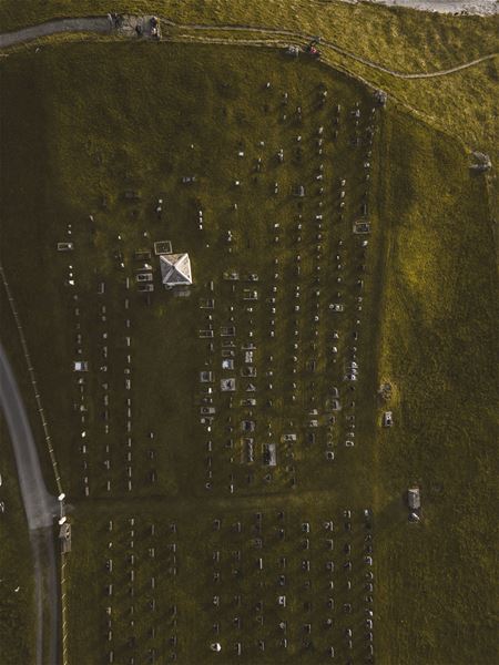 Flyfoto av ein kyrkjegard på grasmark med mange gravsteinar, ei lita bygning og veg langs venstre side..