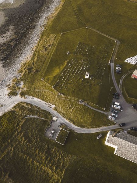 Luftfoto av ei kyrkjegard på grønt jorde ved ei steinete strand, med veg, parkerte bilar og nokre bygg..