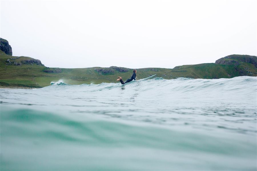 Ein surf ar i våtdrakt ligg på brett i ei bølgje, med grøne klippar og grå himmel i bakgrunnen..