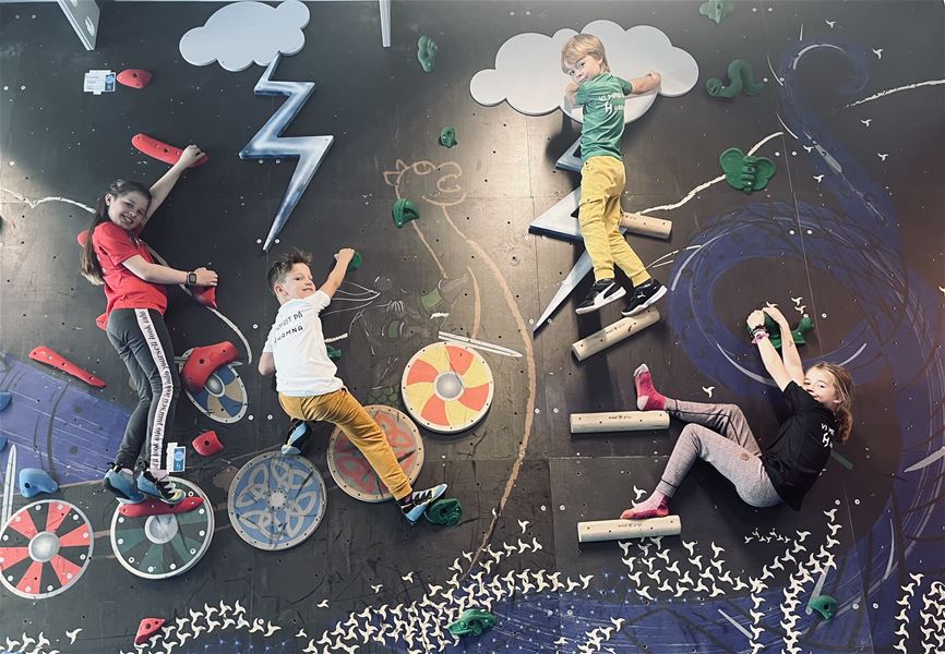 Four children climb a colorful indoor bouldering wall with holds, cloud and lightning shapes, and shield art..