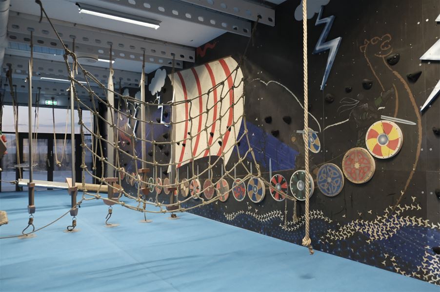 Indoor adventure course with rope net bridge, hanging steps, blue padded floor, and Viking-themed mural wall..