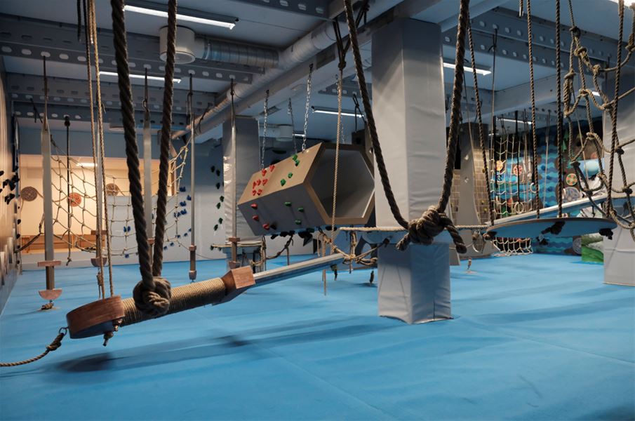 Indoor adventure gym with blue padded floor, hanging ropes and nets, and climbing obstacles under ceiling lights..
