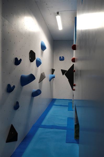 Narrow indoor bouldering corridor with blue climbing holds and volumes on white walls, blue floor mats, and a ceiling light..