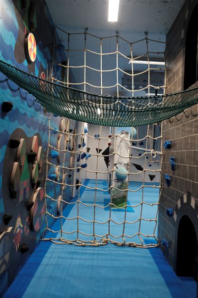 Indoor play area with rope nets, climbing holds on walls, blue padded floor, and a hanging hammock net overhead..