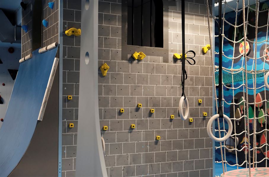 Indoor adventure gym with a gray climbing wall, yellow holds, hanging gymnastic rings, and a rope net..