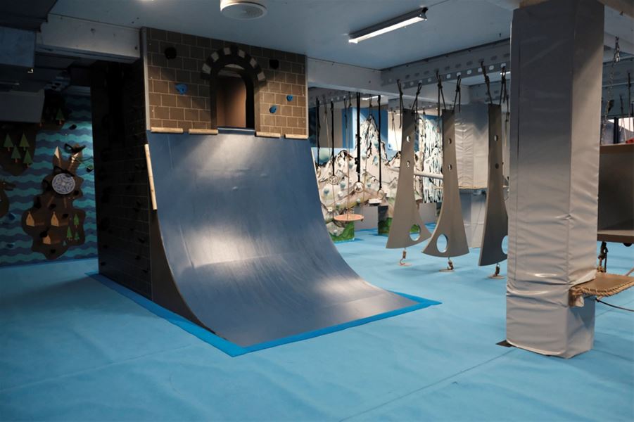 Indoor adventure gym with a curved ramp wall, hanging obstacles, padded columns, and blue safety flooring..