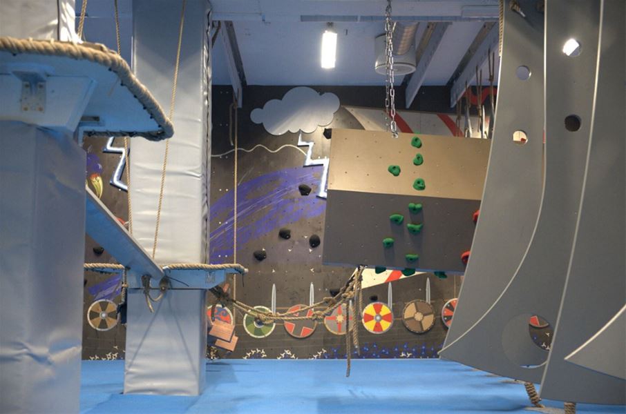 Indoor obstacle course with padded platforms, ropes, and a small climbing wall with green holds in a gym..