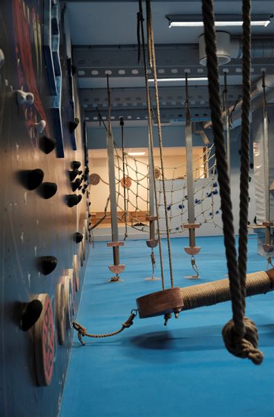 Empty indoor adventure gym with climbing holds, hanging ropes, net obstacles, and a padded blue floor..