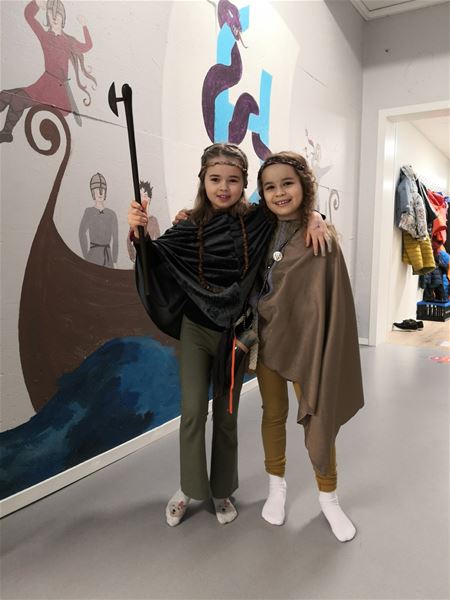 Two people in cloak costumes pose with arms around each other beside a Viking ship wall mural in a hallway..