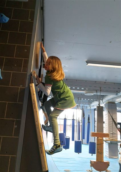 A child climbs an indoor climbing wall in a gym, reaching for a wooden hold with hanging obstacles behind..