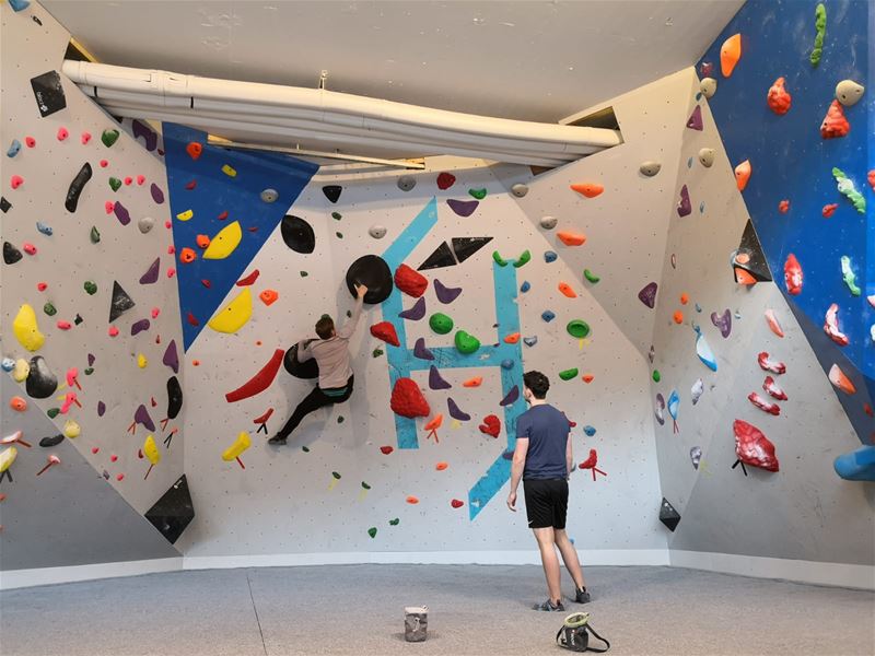 Two people in a bouldering gym; one climbs on a colorful wall of holds while another stands watching below..