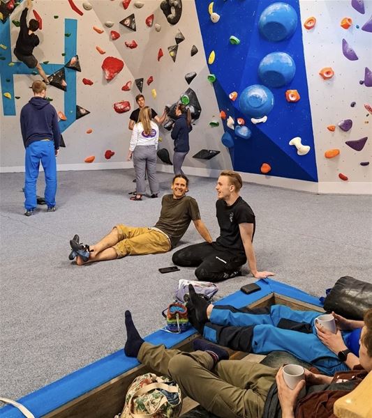 People rest and chat on padded flooring inside an indoor bouldering gym with colorful climbing holds on walls..