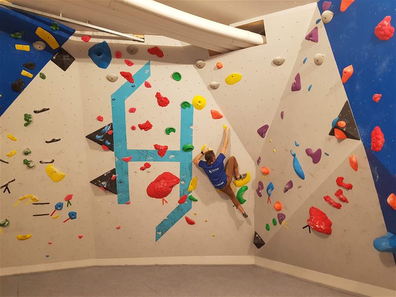 A climber in a blue shirt ascends an indoor bouldering wall with colorful holds on angled panels under ceiling lights..