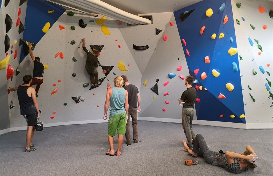 Indoor bouldering wall with colorful holds as several climbers practice, watch, and rest on padded floor..