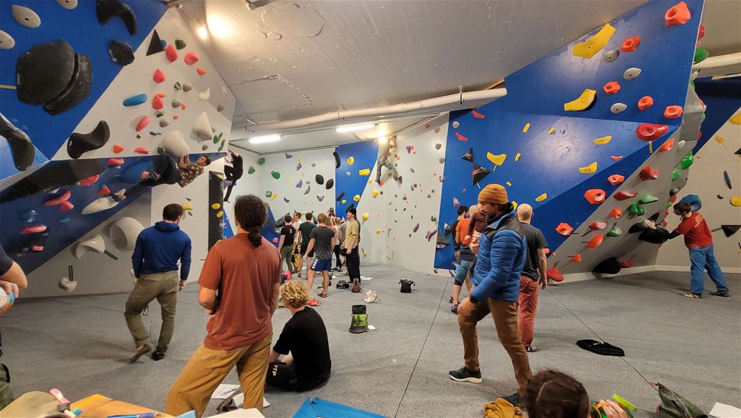 Crowded indoor bouldering gym with climbers and spectators under blue walls covered in colorful holds and volumes..