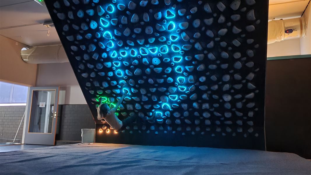 Indoor climber reaches on an overhanging wall with many holds, illuminated by blue and green LED lights above mats..
