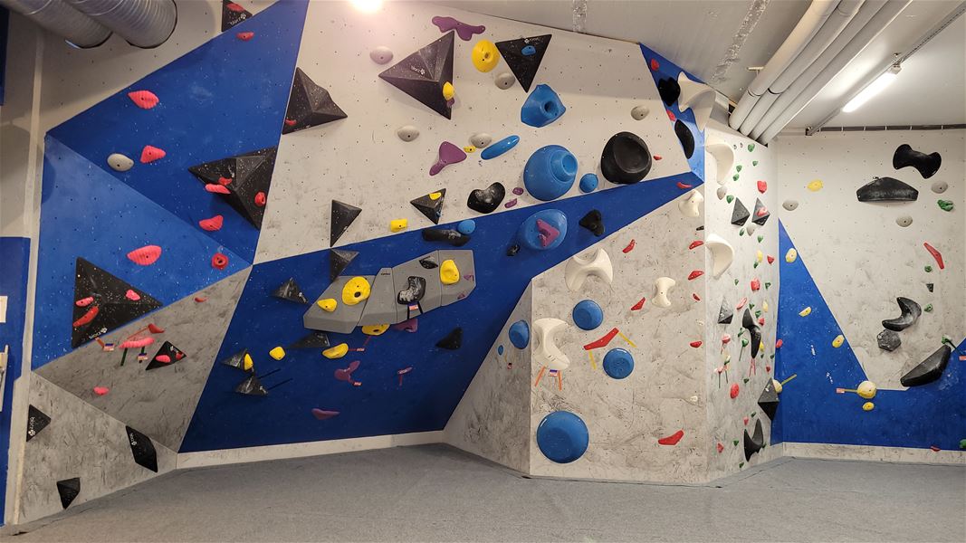 Indoor bouldering wall with blue and gray panels, assorted climbing holds and volumes, and padded floor below..