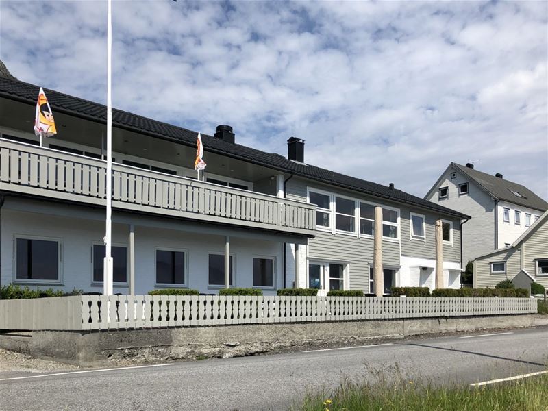 Two-story gray building with balcony, flags and fenced yard beside a paved road under a partly cloudy sky..