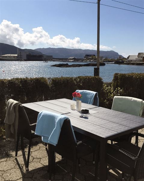 Outdoor patio table with chairs and draped blankets, a vase of flowers, and a sunny harbor with mountains beyond..