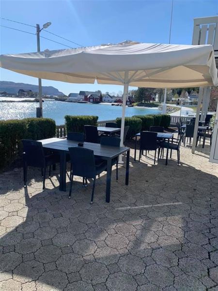 Outdoor patio with black tables and chairs under a large umbrella beside a calm waterfront and distant houses..