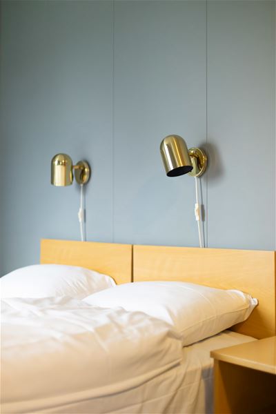 White bed with pillows and wooden headboard beneath two brass wall lamps on a light blue wall in a calm room..